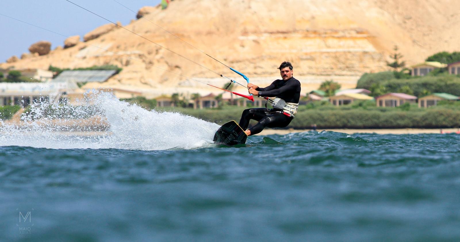 Kitesurfista girando sobre el agua y levantando una estela junto a una costa rocosa.