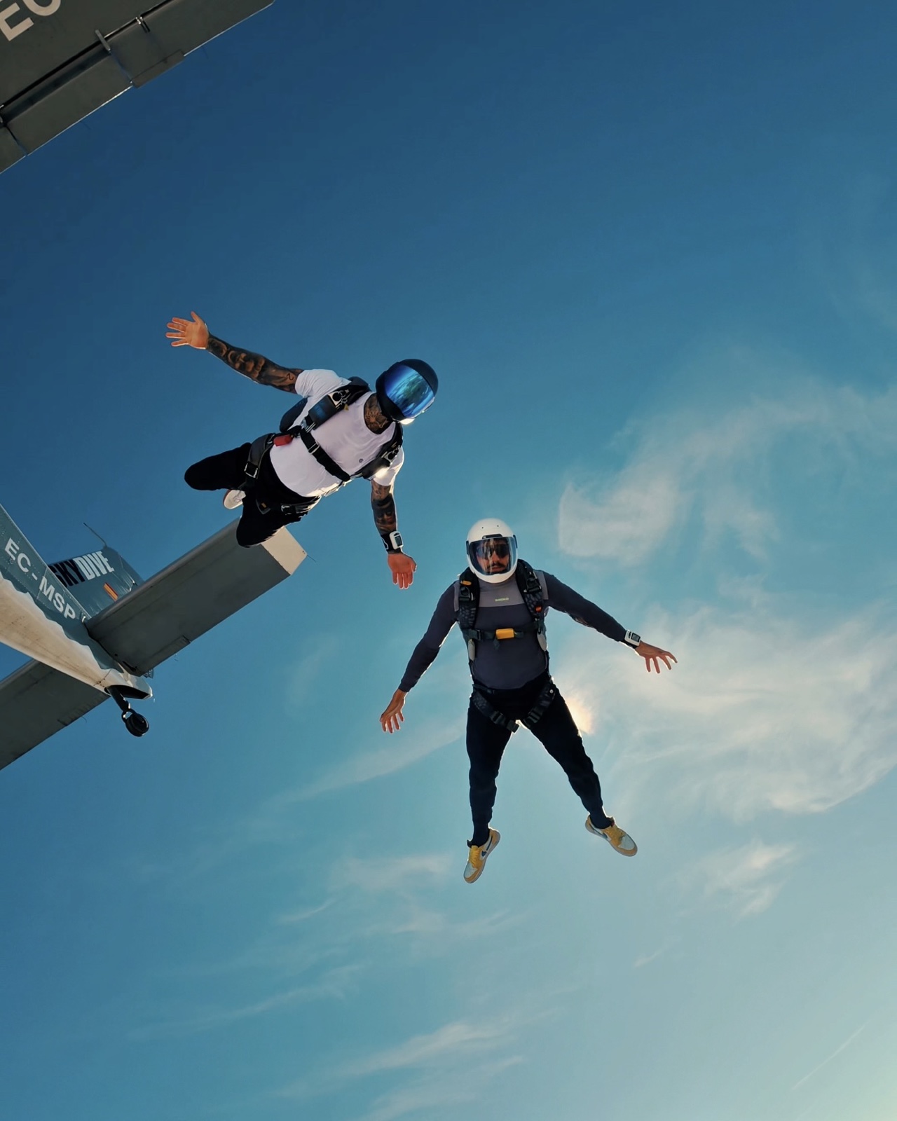 Dos paracaidistas en el aire junto a un avión, sobre un fondo de cielo azul.