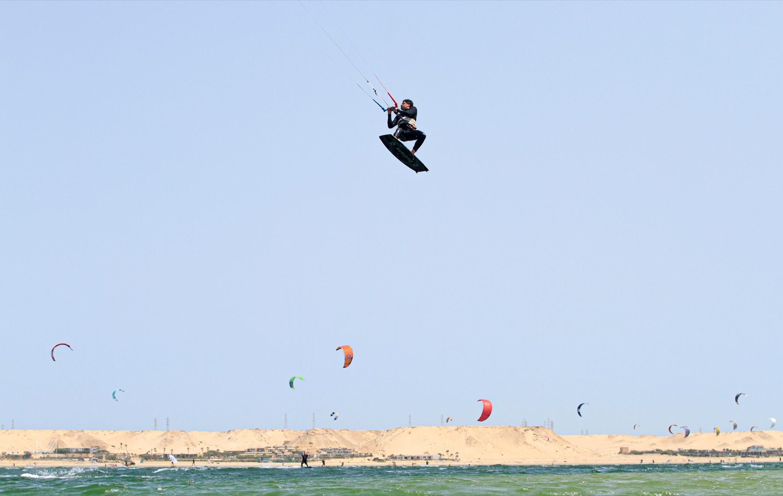 Kitesurfista en un salto alto sobre el mar, con otras cometas junto a la costa al fondo.