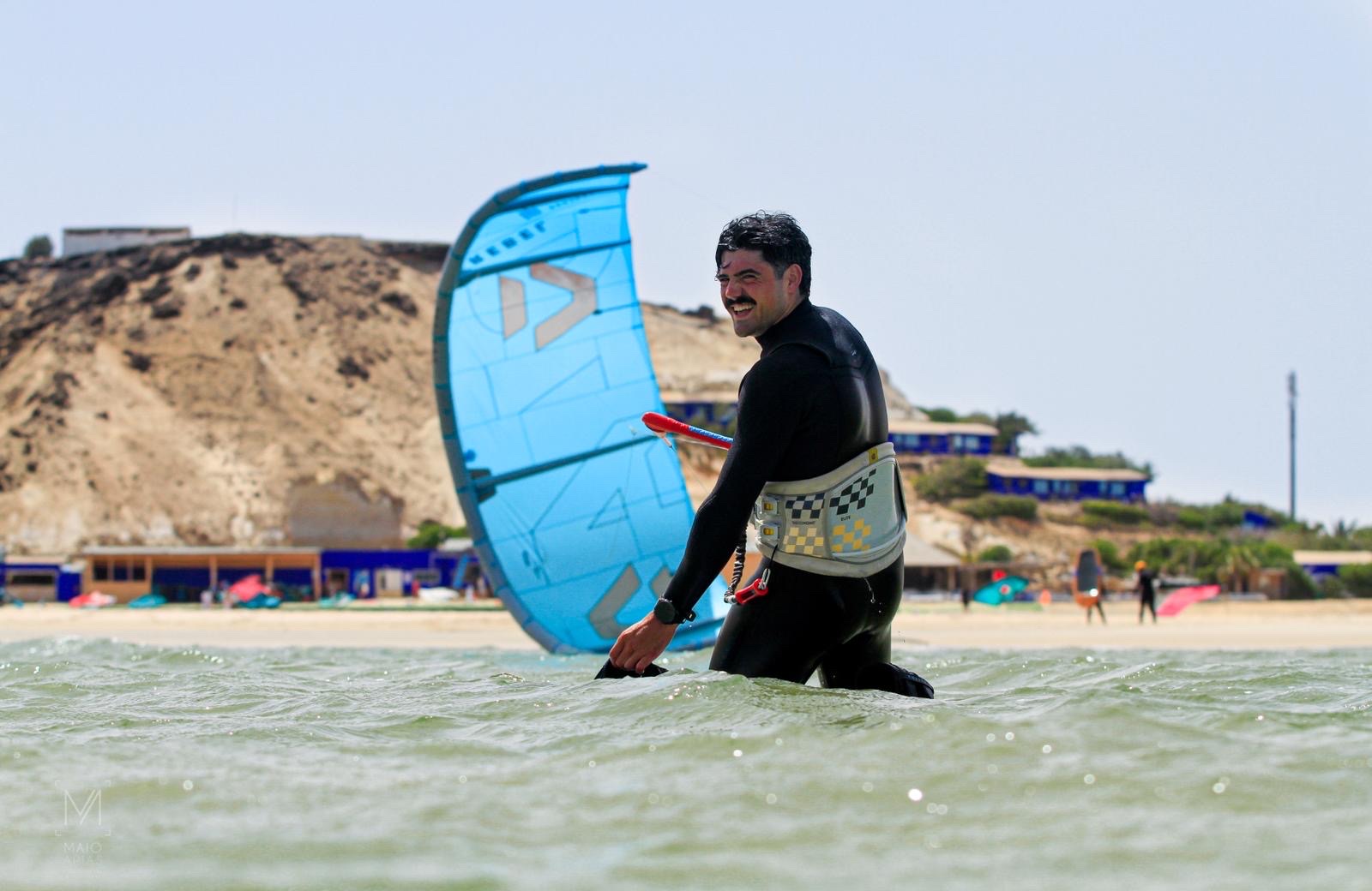 Deportista con neopreno y arnés en el agua, junto a una cometa azul y una playa rocosa.