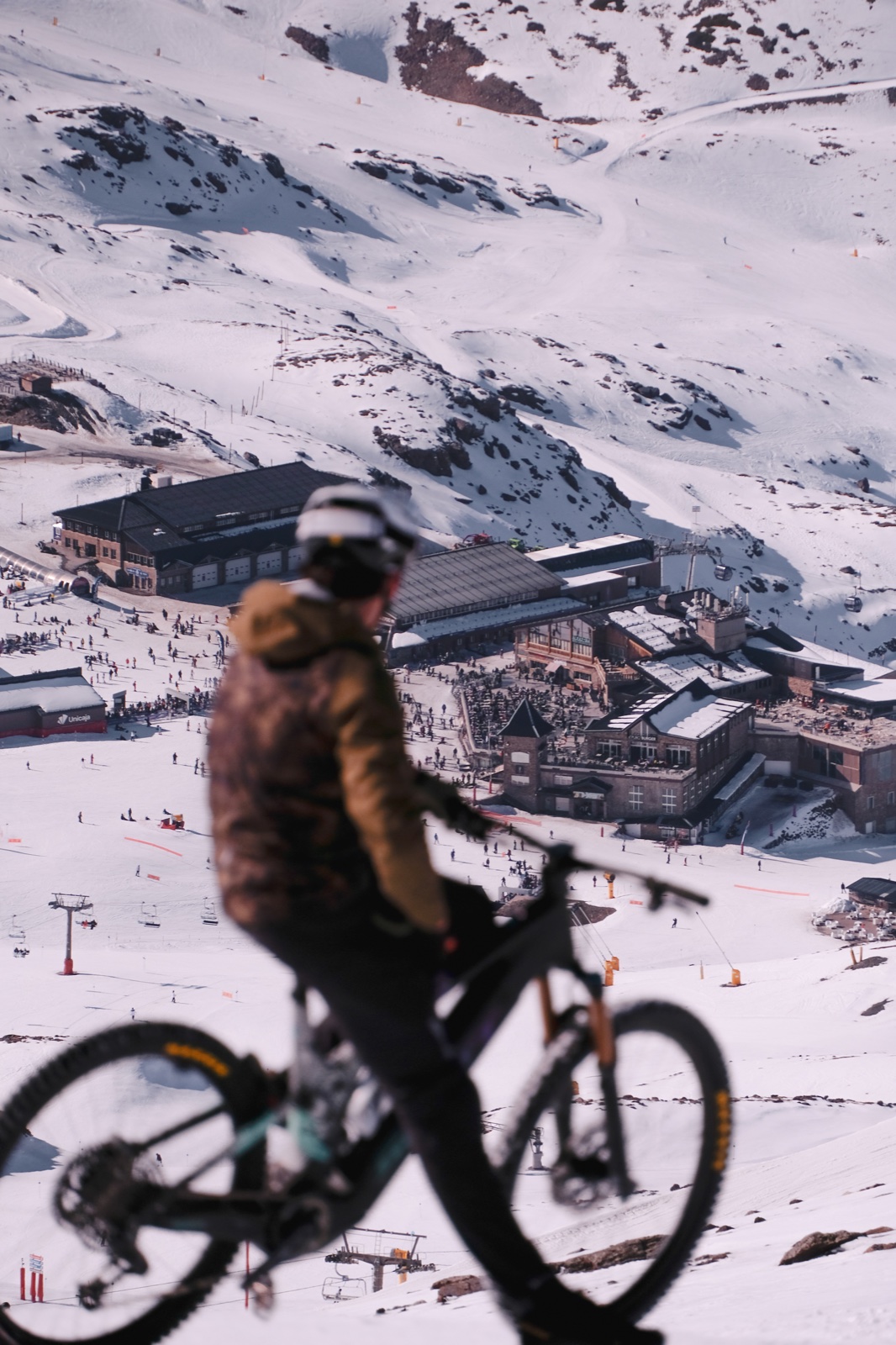Ciclista con casco junto a una bicicleta en una ladera nevada, con edificios al fondo.