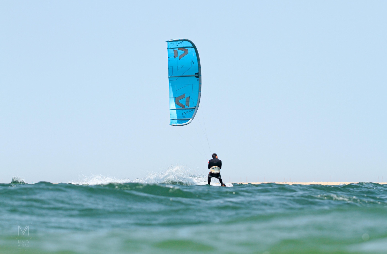 Kitesurfista con cometa azul navegando entre olas bajo un cielo despejado.