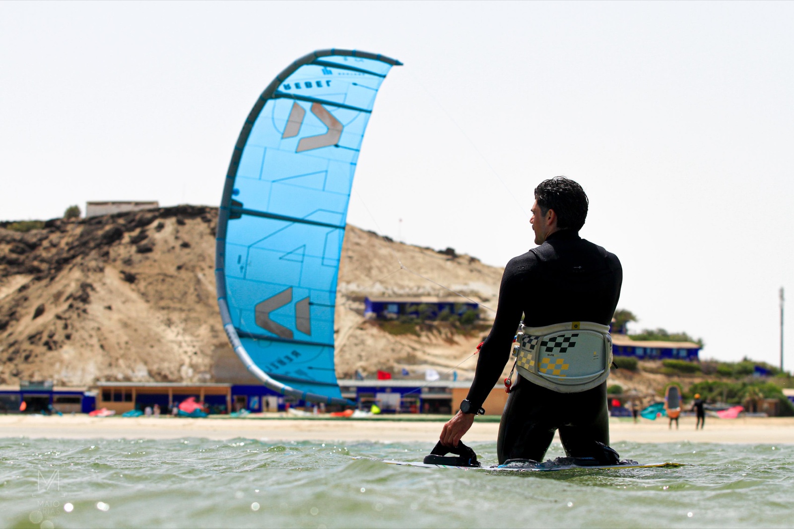 Deportista de espaldas, con neopreno y arnés, junto a una cometa azul en la orilla.
