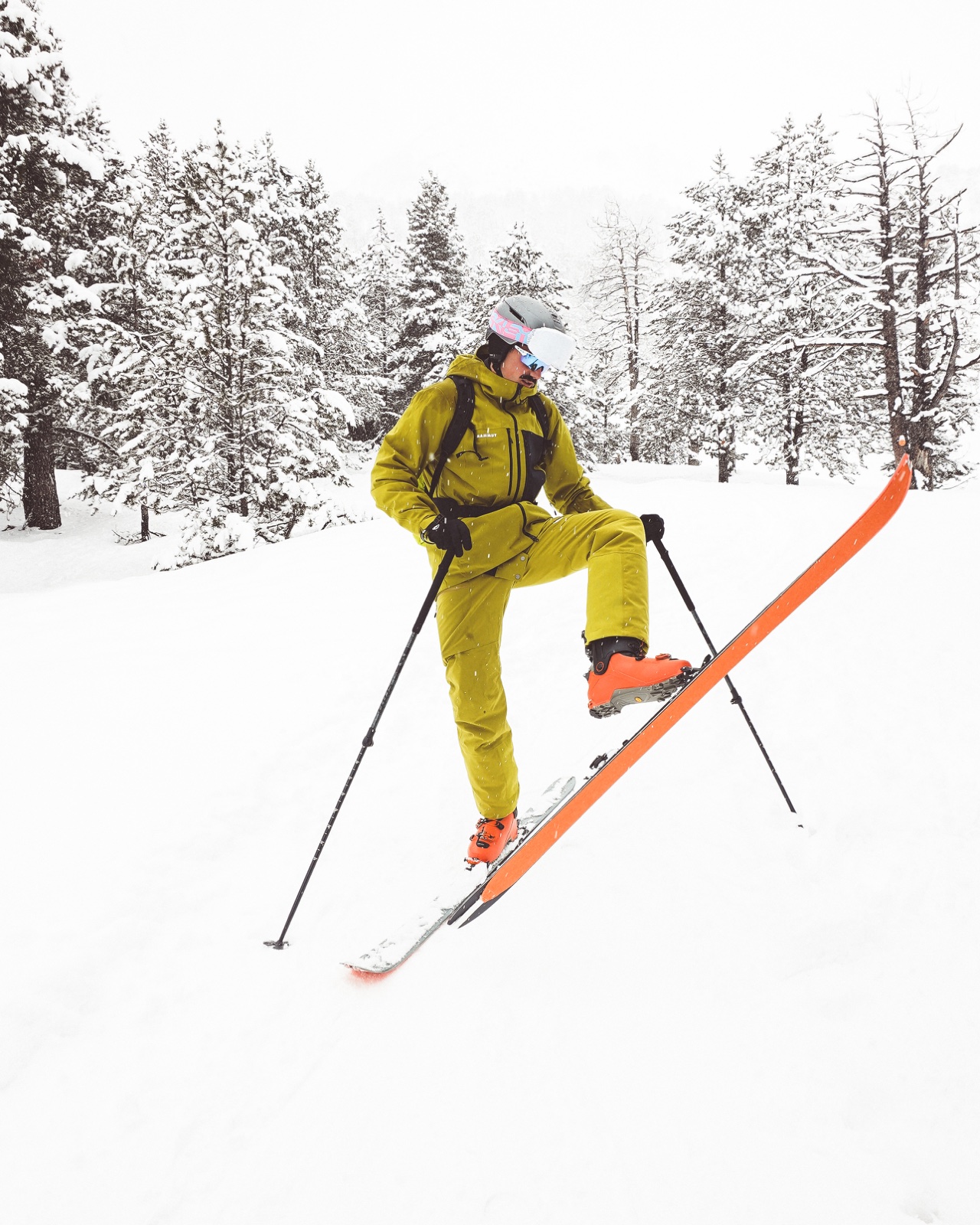 Esquiador vestido de amarillo levantando un esquí naranja en un paisaje de pinos nevados.