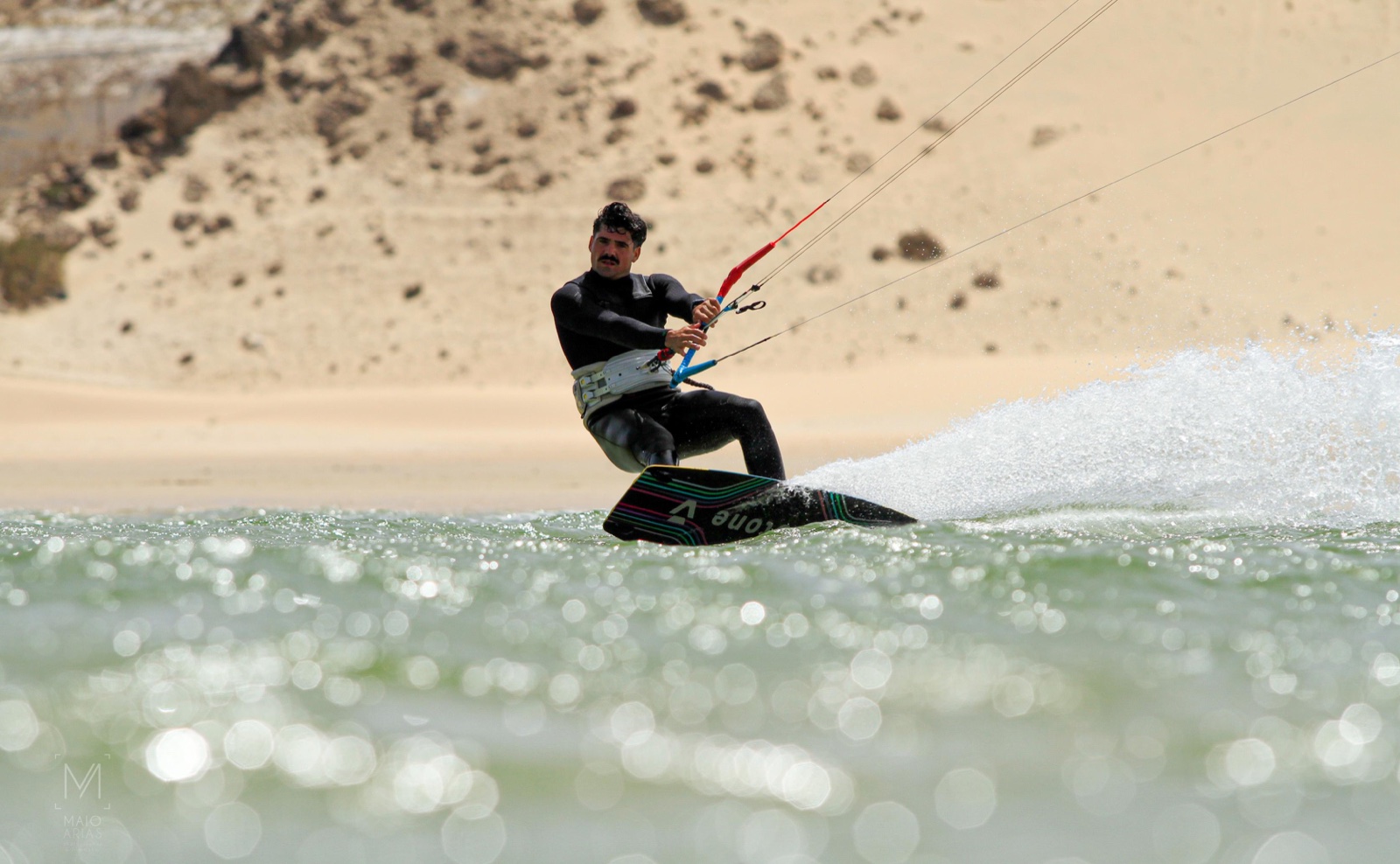 Kitesurfista trazando un giro y levantando agua frente a una playa de arena y roca.
