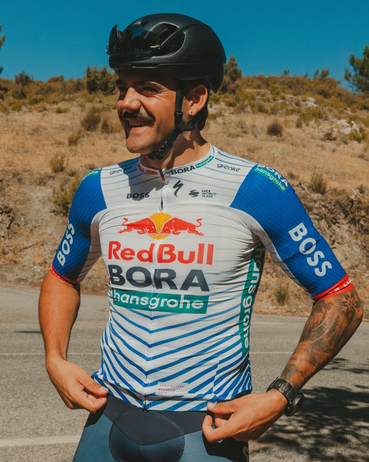 Ciclista con casco negro y maillot azul y blanco sonriendo en un camino junto a una ladera seca.