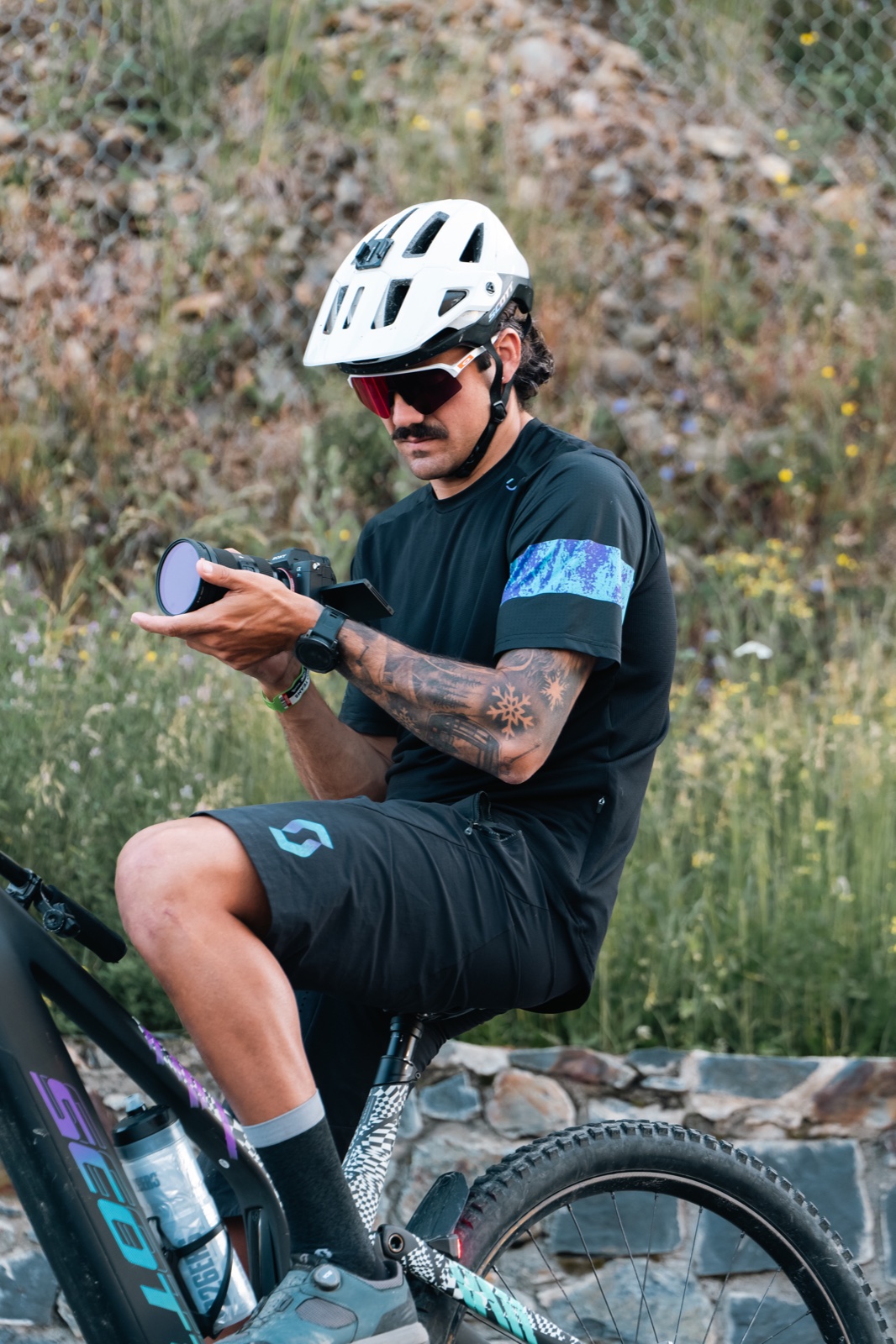 Ciclista con casco blanco y ropa negra sentado en una bicicleta, mirando un objeto que sostiene entre las manos.