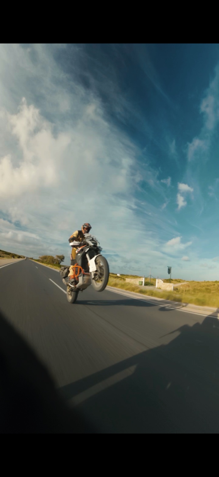 Motorista levantando la rueda delantera de una moto en una carretera, bajo un cielo con nubes.