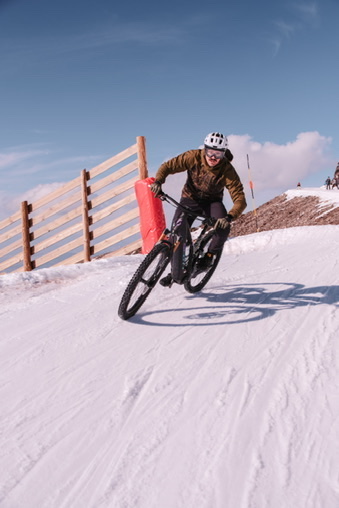 Ciclista con casco inclinando una bicicleta sobre una pista nevada junto a una valla de madera.