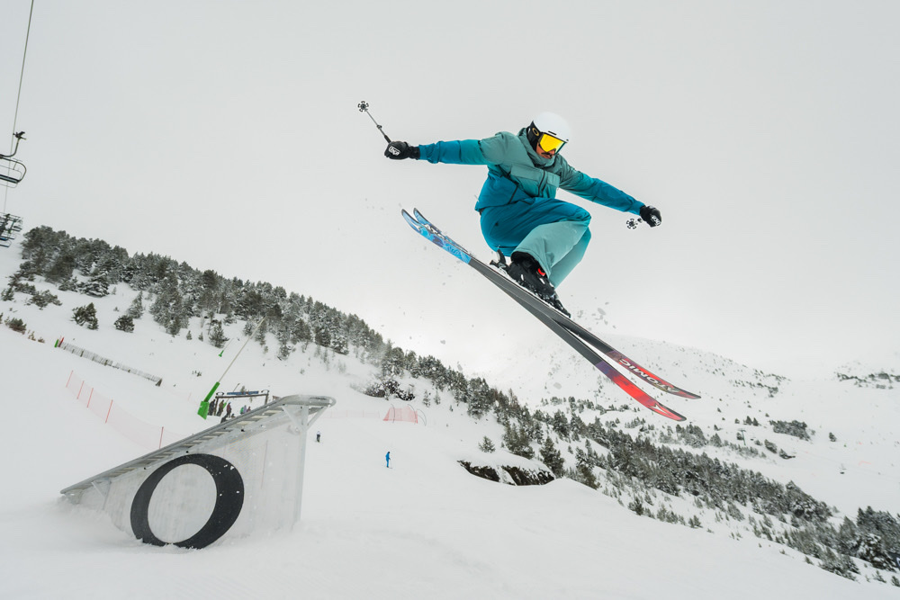 Esquiador con ropa azul saltando junto a un módulo de snowpark en una estación de montaña.