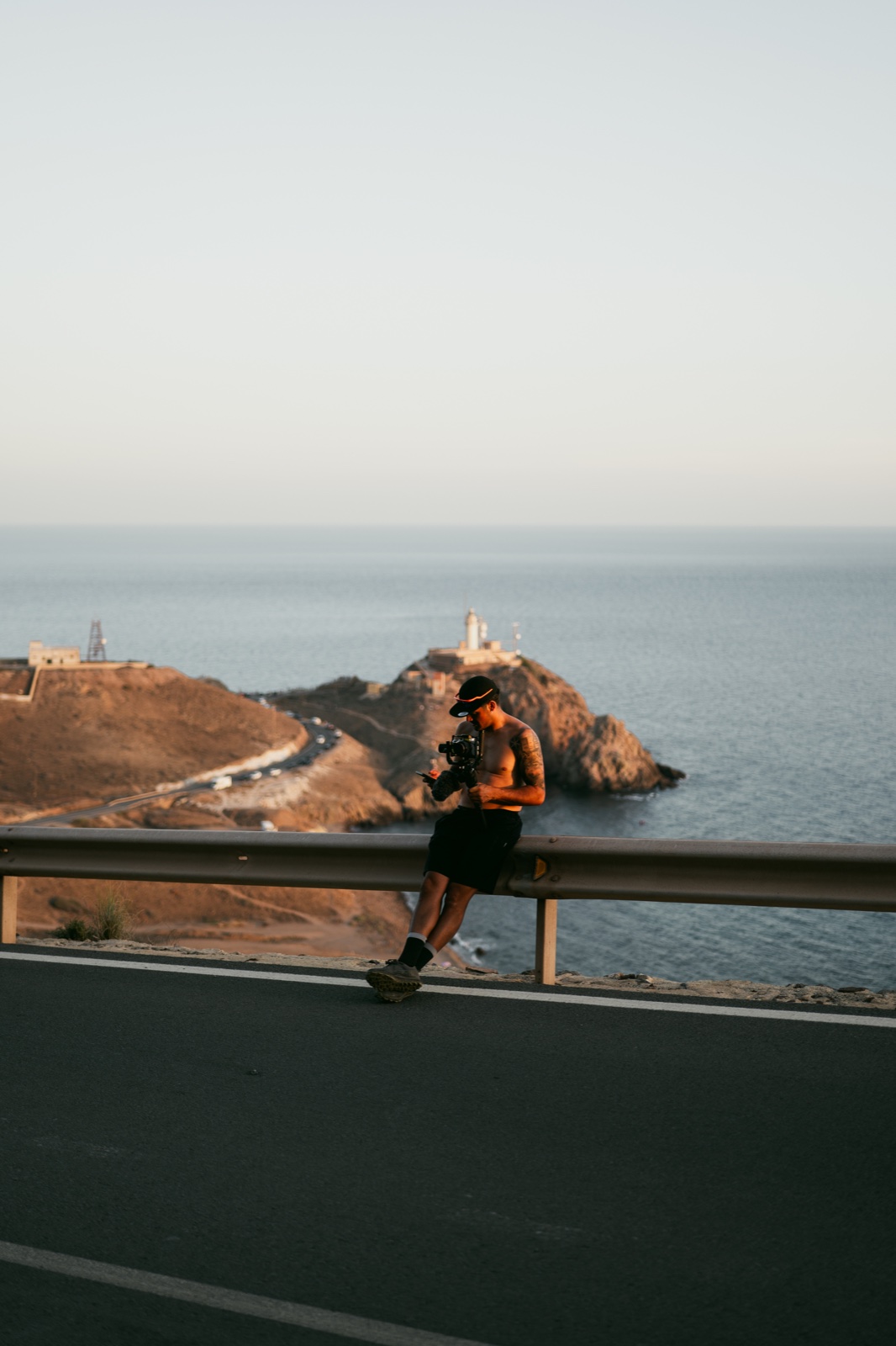 Hombre sentado en una barrera de carretera junto a una costa rocosa y el mar al atardecer.