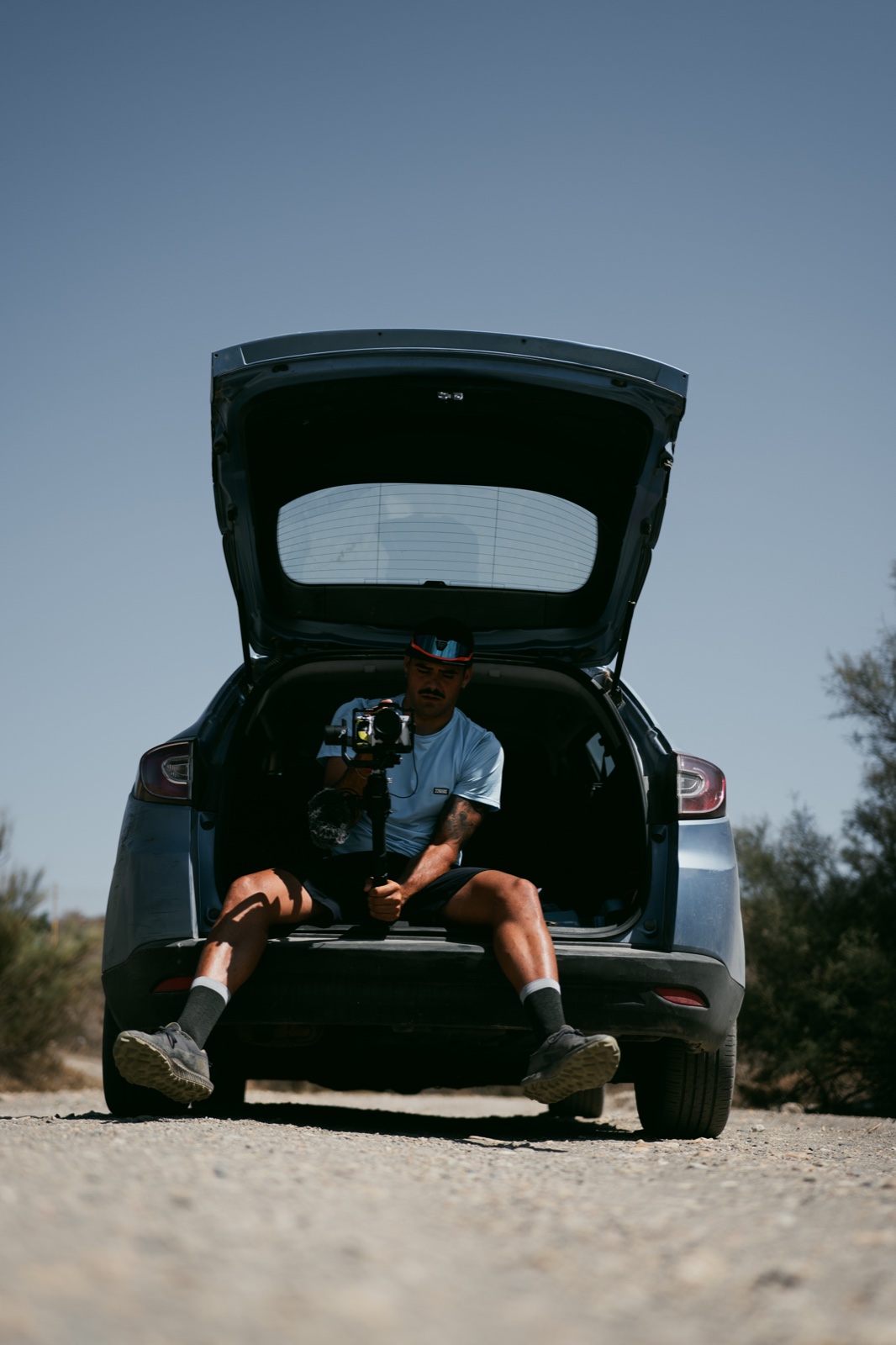 Hombre sentado en el maletero abierto de un coche aparcado al aire libre bajo un cielo azul.