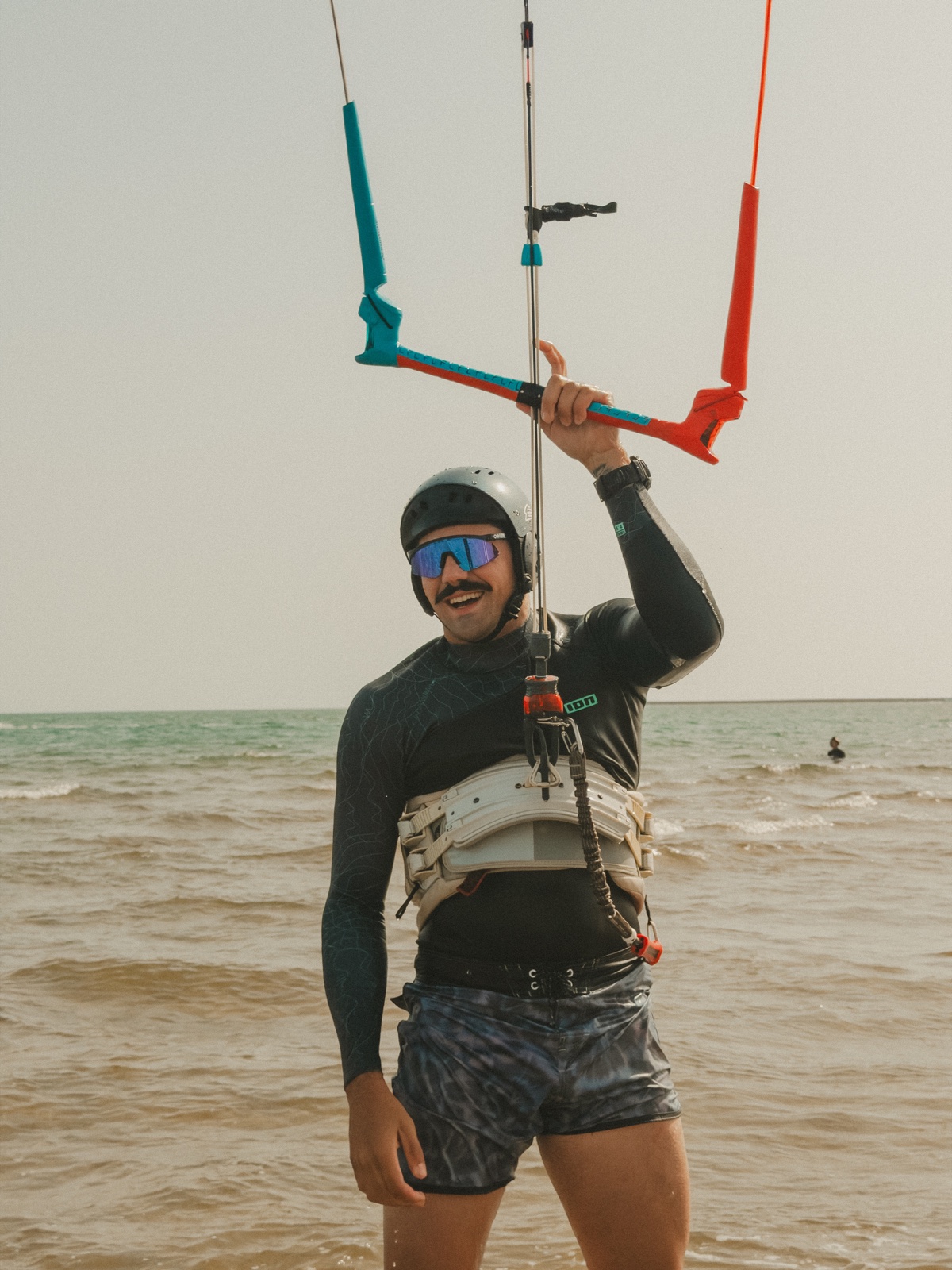 Deportista con casco, gafas y neopreno sujetando una barra de kitesurf en la orilla del mar.