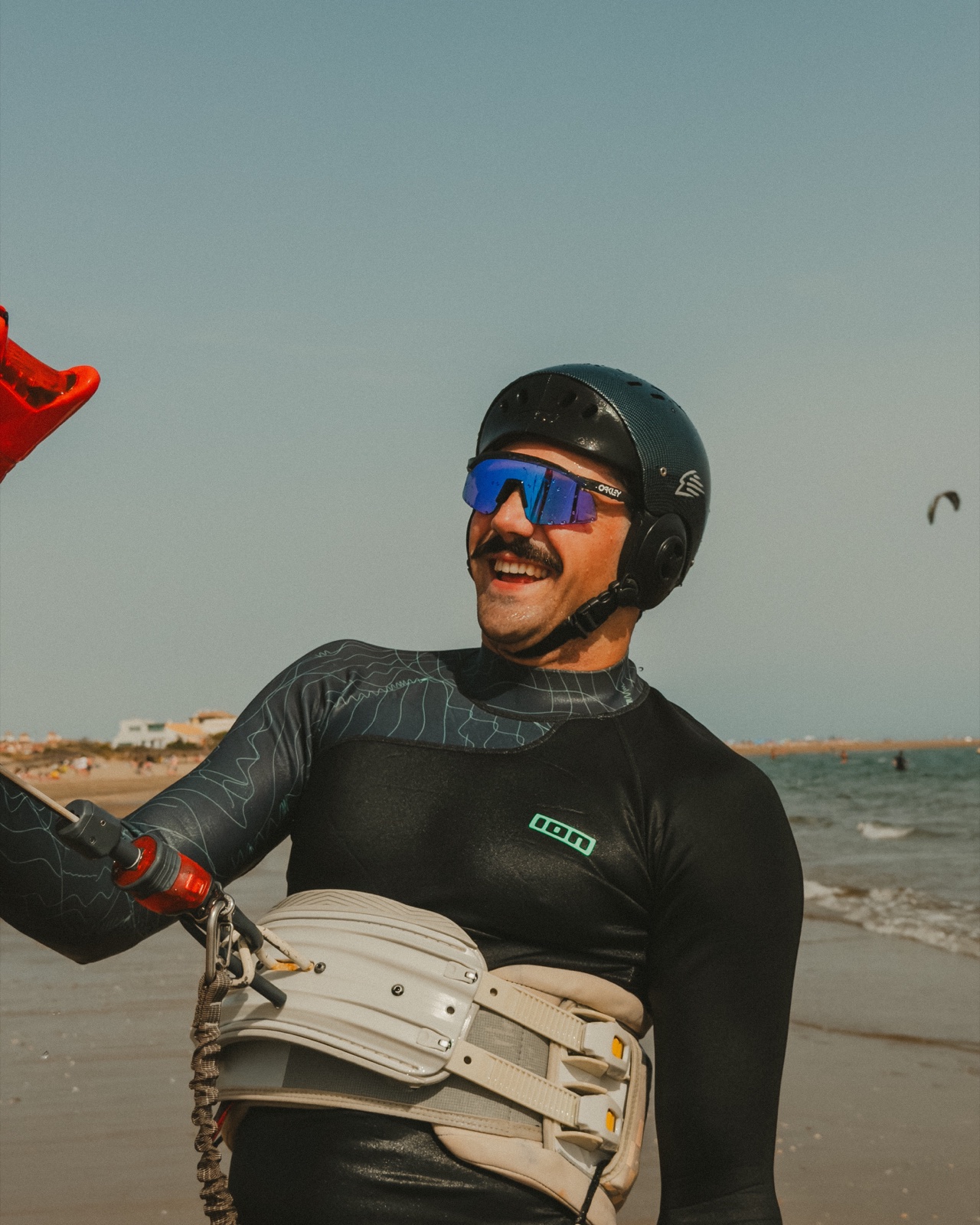Deportista con casco negro, gafas azules, neopreno y arnés sonriendo junto al mar.