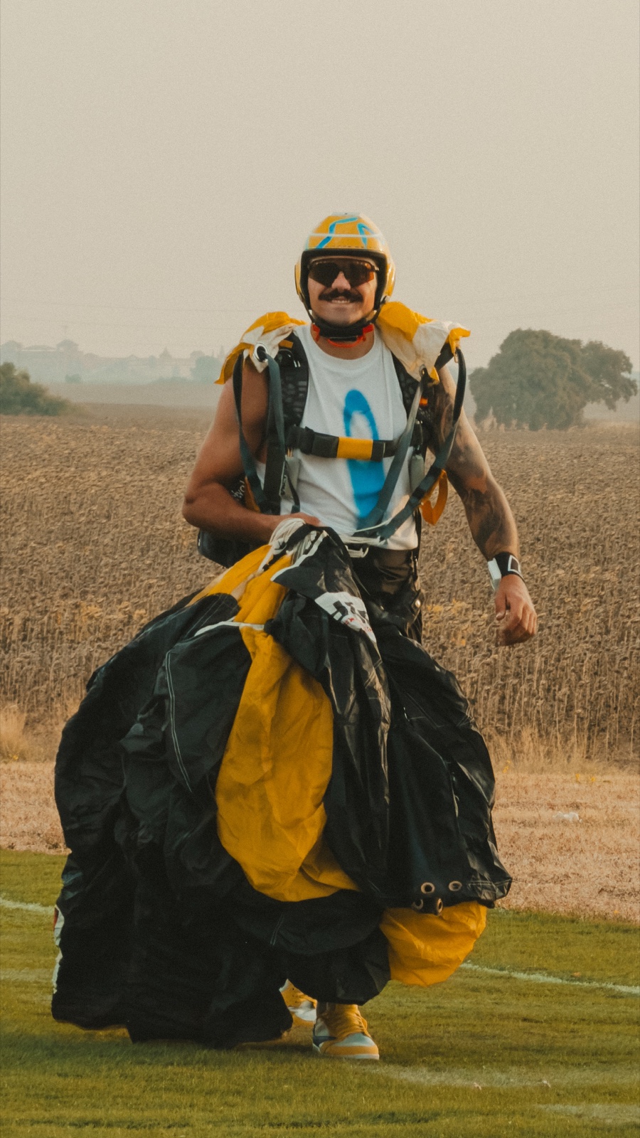Paracaidista con casco amarillo y azul caminando por el campo con el paracaídas recogido en los brazos.