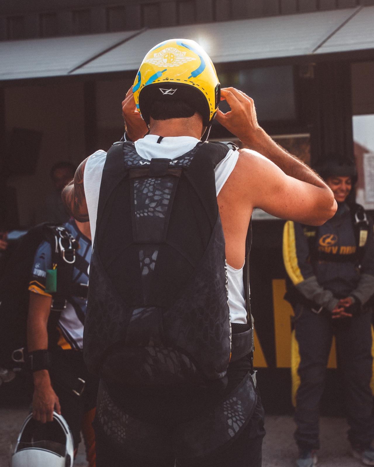 Paracaidista de espaldas ajustándose un casco amarillo y azul, con arnés y mochila junto a un hangar.
