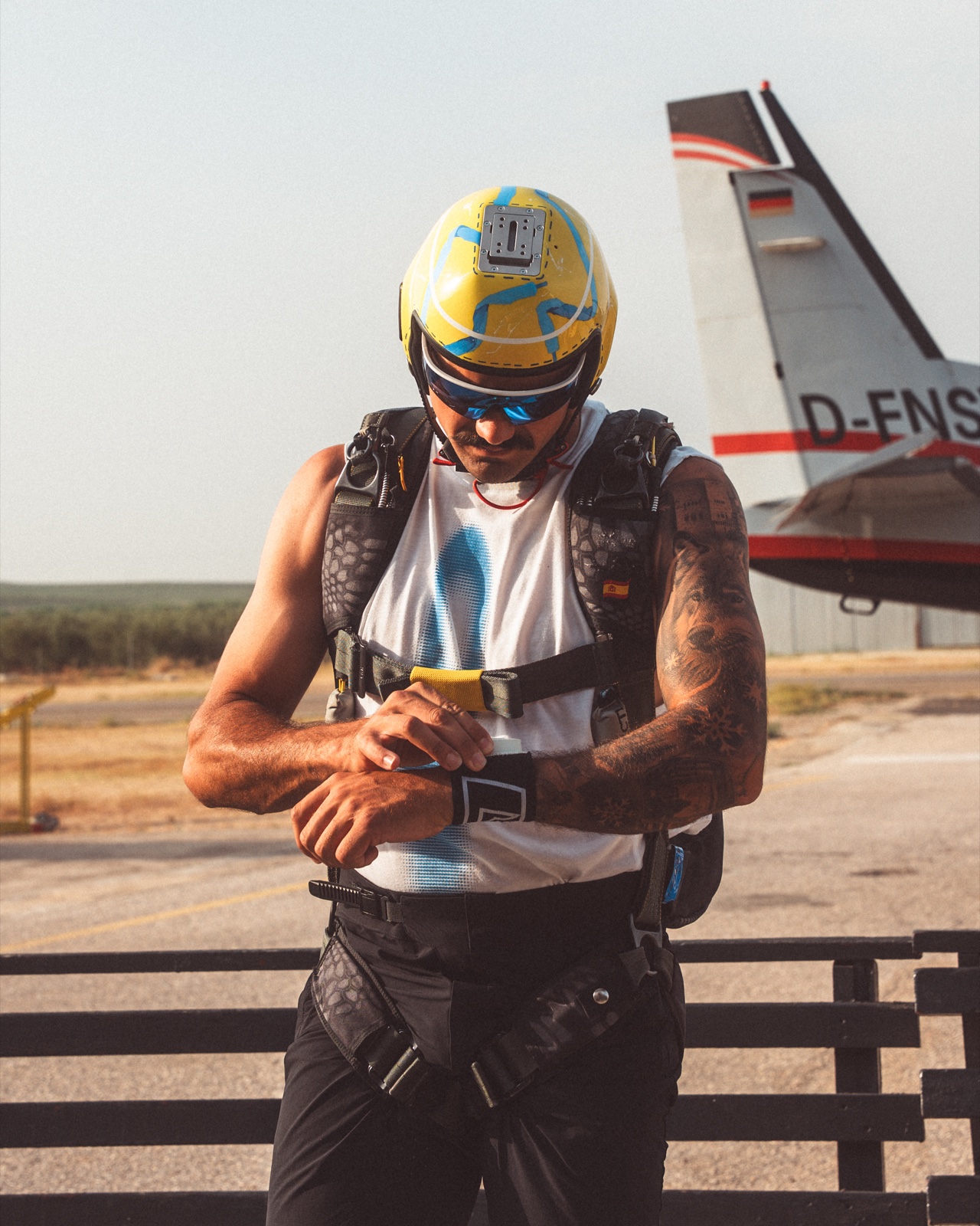 Paracaidista con casco amarillo y azul revisando un dispositivo en la muñeca junto a un avión.