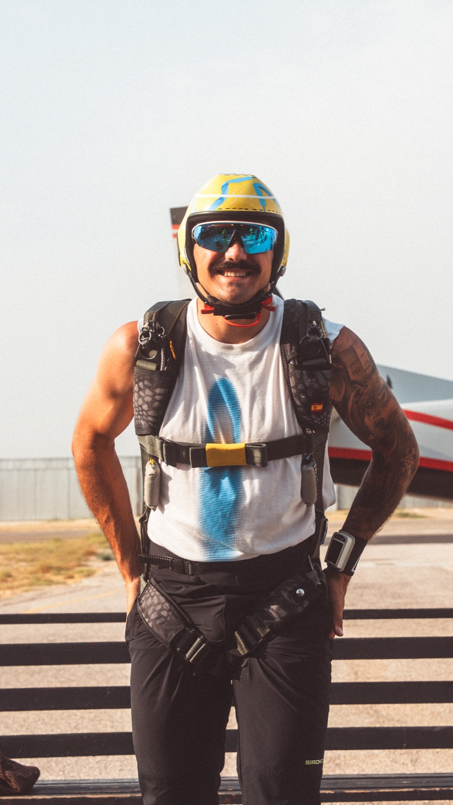 Paracaidista sonriendo con casco amarillo y azul, gafas y arnés, junto a una valla del aeródromo.