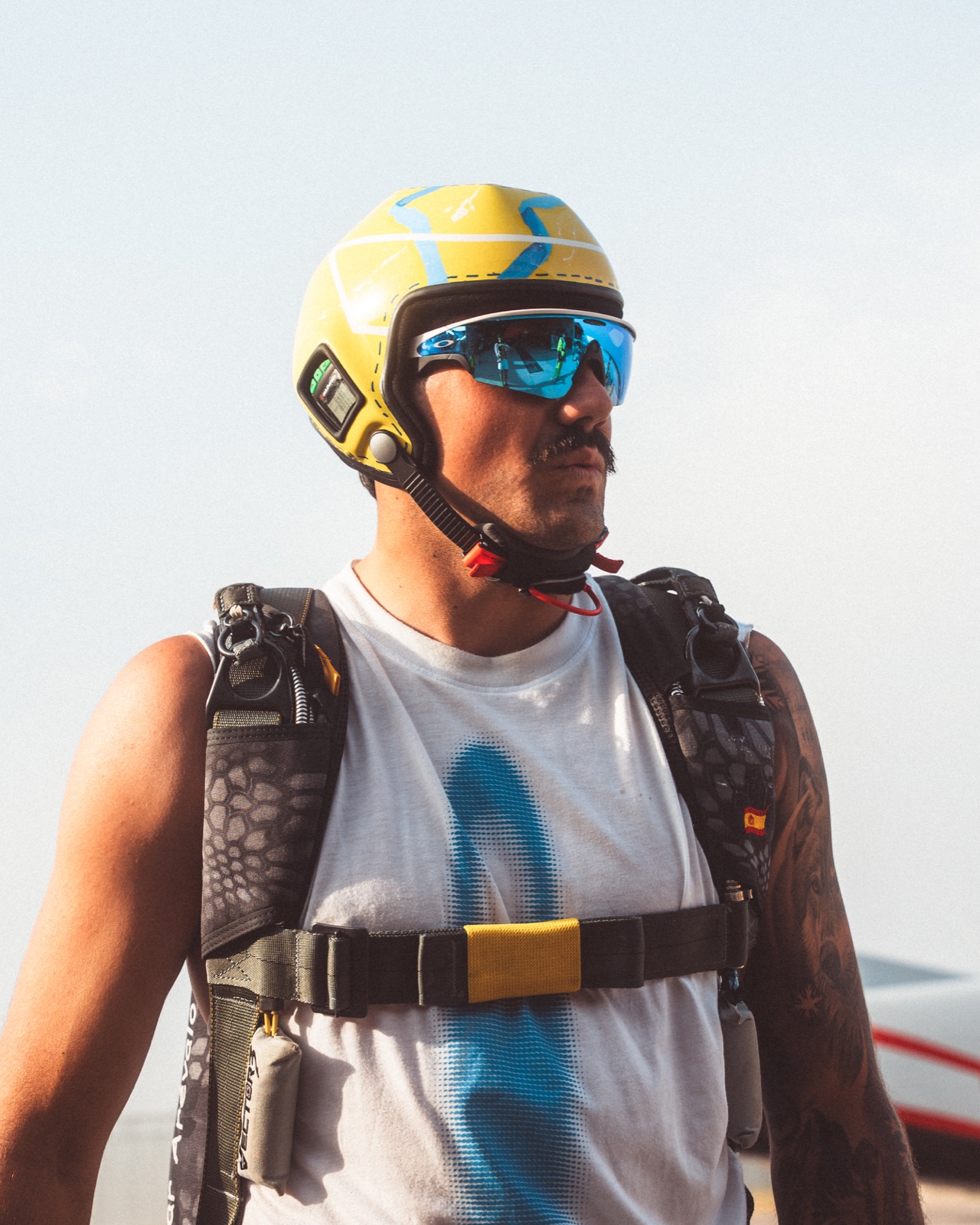 Retrato lateral de un paracaidista con casco amarillo y azul, gafas y arnés bajo un cielo claro.
