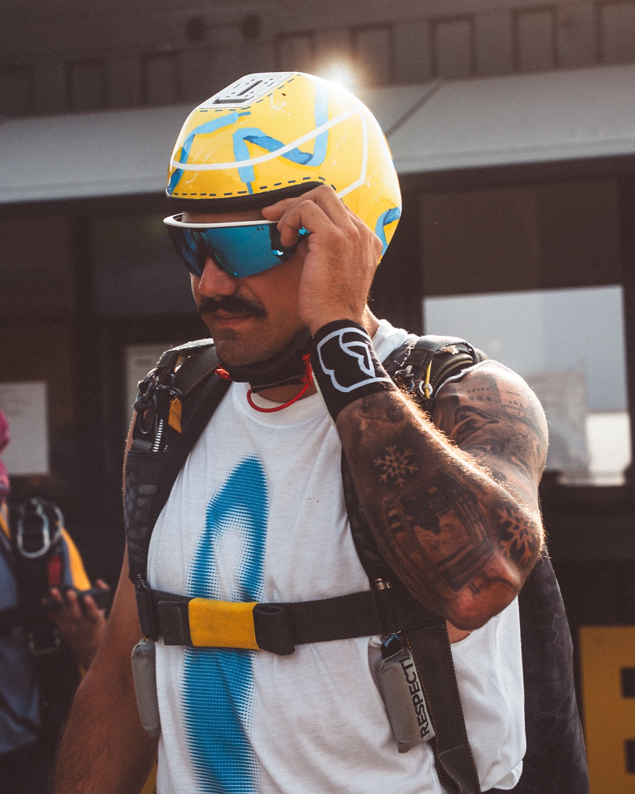 Paracaidista con casco amarillo y azul ajustándose las gafas junto a un hangar.