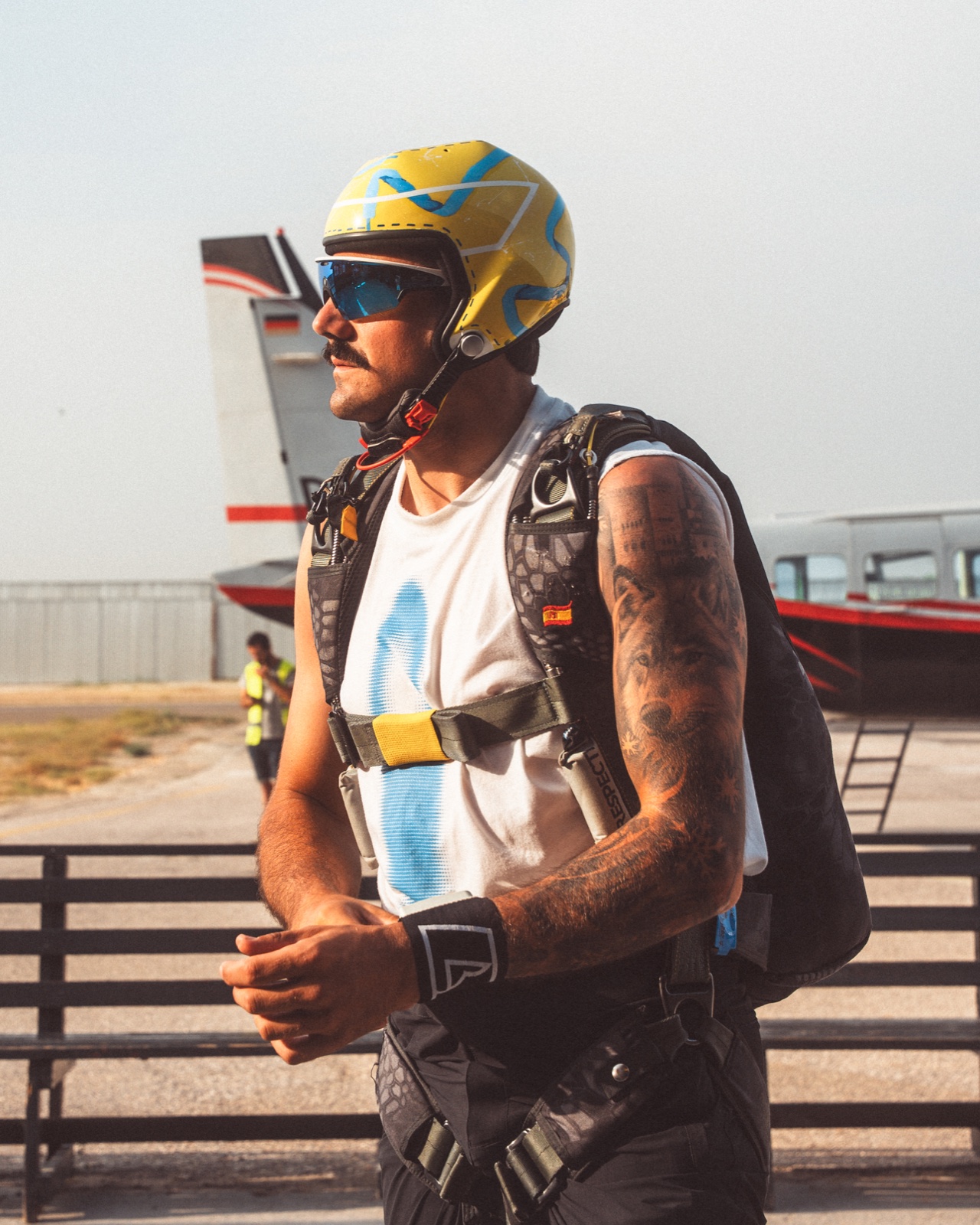 Paracaidista con casco amarillo y azul y arnés mirando hacia un lado junto a un avión.