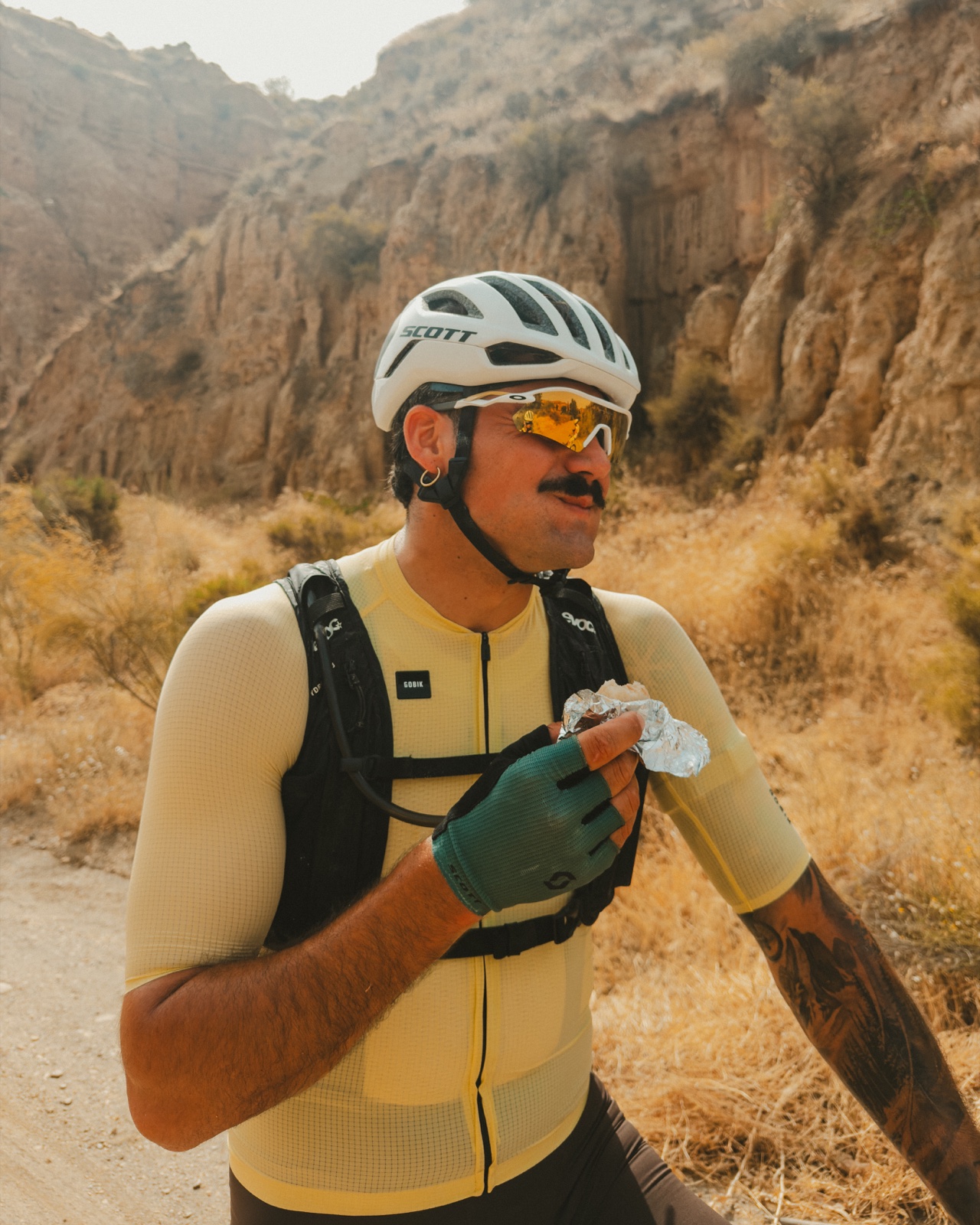 Ciclista con casco blanco y maillot amarillo sosteniendo comida durante una pausa en un paisaje de roca y monte.
