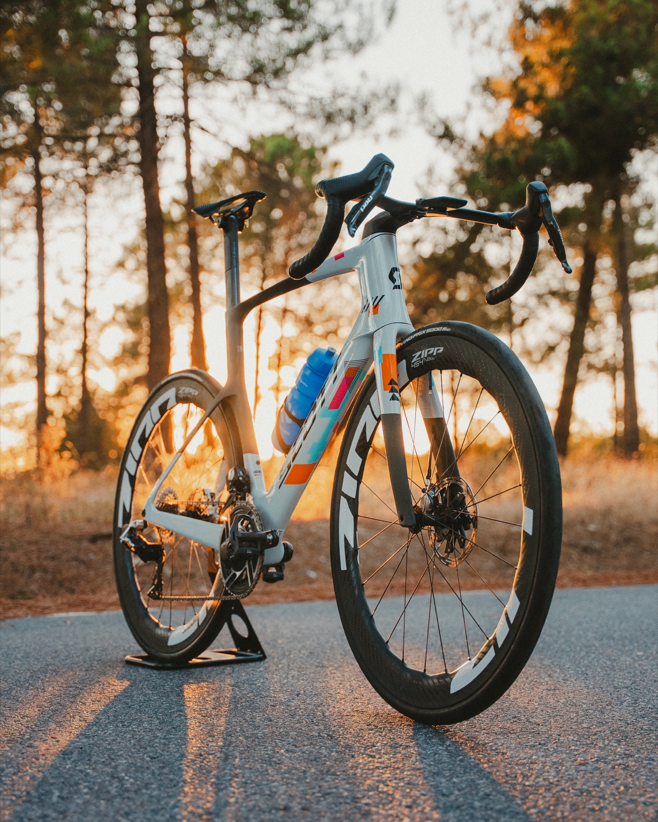 Bicicleta de carretera azul y naranja fotografiada sobre el asfalto entre pinos con luz de atardecer.