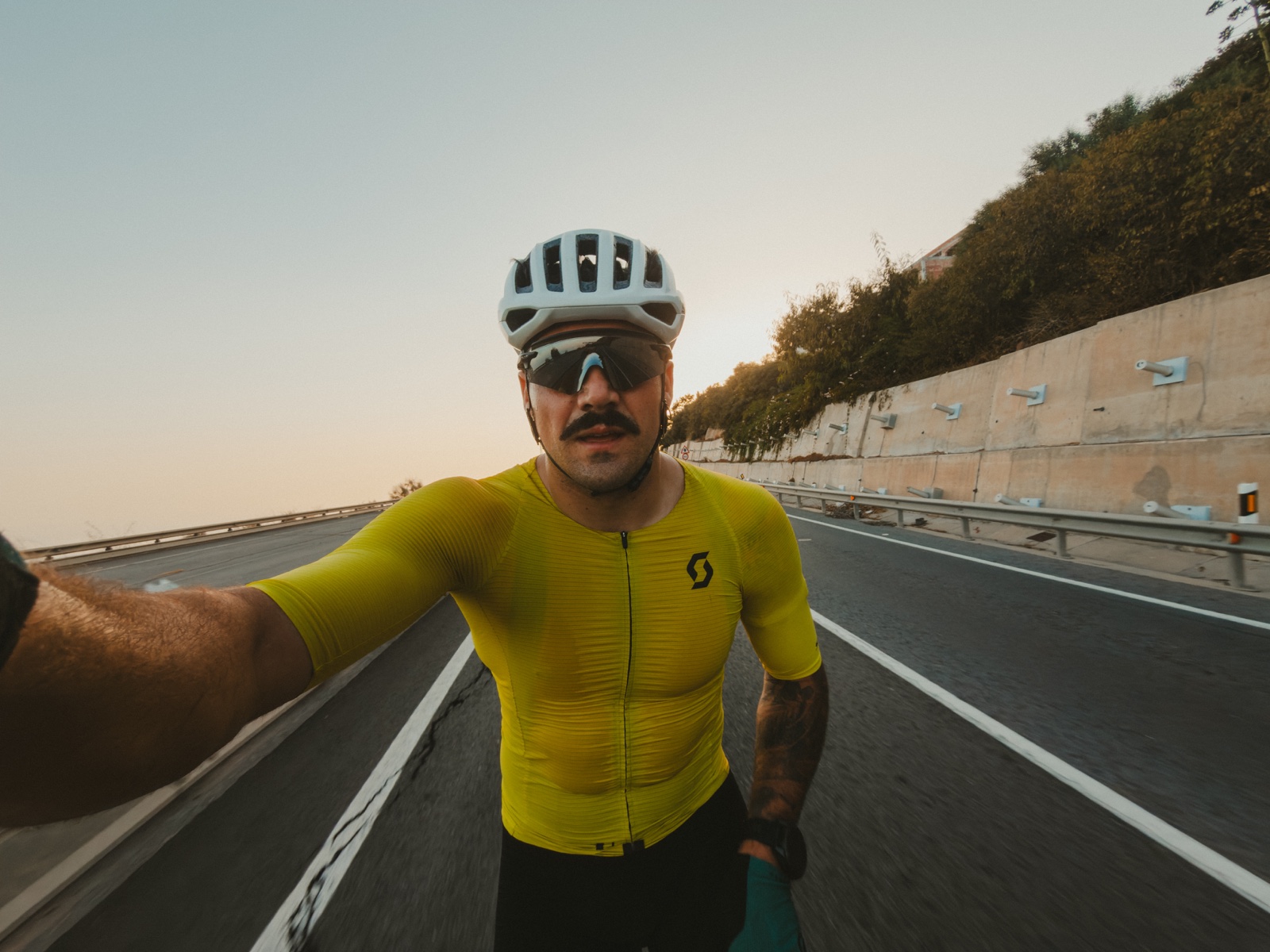 Ciclista con casco blanco y maillot amarillo fotografiado de cerca en una carretera vacía, con el brazo extendido hacia la cámara.